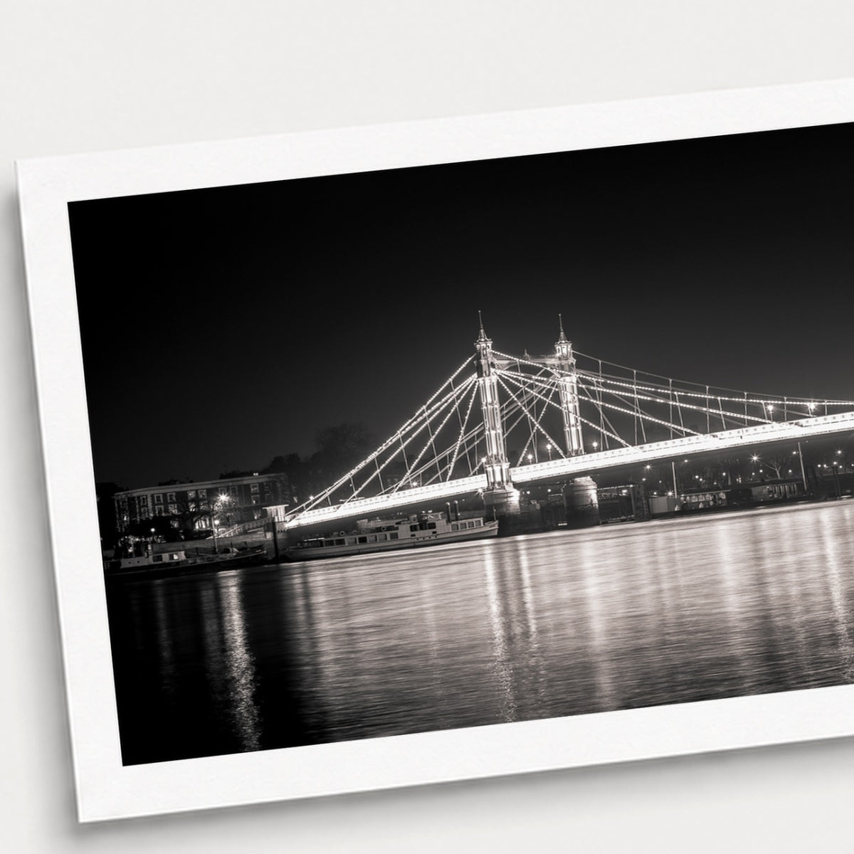 A photographic print of Albert Bridge by Night, shown with a white border and angled presentation on a light background.