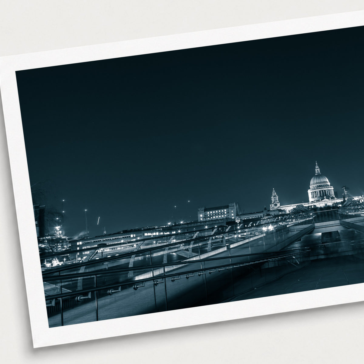 A black and white photographic print of Millennium Bridge, shown with a white border and angled presentation on a light background.